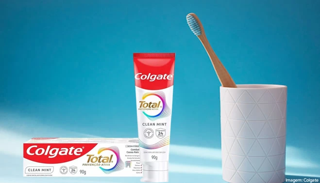 Um tubo de pasta de dente Colgate Total Clean Mint vermelho e branco e sua caixa correspondente estão em pé sobre uma superfície azul clara. À direita, uma escova de dentes de bambu está em um suporte cilíndrico branco. O texto "Imagem: Colgate" está no canto inferior direito.