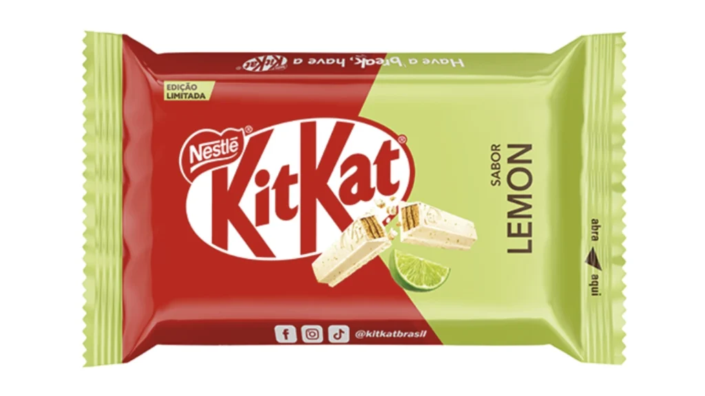 Uma embalagem de Kit Kat de edição limitada, com um design dividido verticalmente em vermelho e verde claro. No lado vermelho, o logotipo "Kit Kat" em letras brancas com contorno vermelho é proeminente, e acima dele, em um círculo branco, está o logotipo da "Nestlé". Uma fatia de chocolate branco Kit Kat com recheio de wafer e uma fatia de limão verde fresco são mostradas. A parte superior da embalagem possui um texto verde claro que, quando virado, diz "Have a break, have a Kit Kat". No lado verde claro, a palavra "LEMON" (limão) está escrita verticalmente em letras verdes escuras, com a palavra "SABOR" (sabor) acima dela. No canto inferior direito, há uma seta preta apontando para baixo com as palavras "abra aqui" em letras verdes. Na parte inferior da embalagem, em um fundo vermelho, estão ícones de redes sociais (Facebook, Instagram, TikTok) e o nome de usuário "@kitkatbrasil".