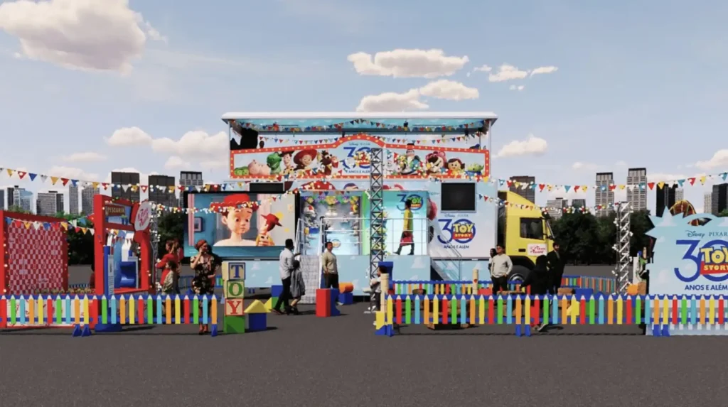 Uma imagem mostra uma montagem ao ar livre do "Toy Story - 30 Anos e Além", um evento gratuito para crianças. Ao centro, um caminhão azul e branco foi transformado em um palco móvel com telas mostrando personagens de Toy Story e a marca do evento. Ao redor do caminhão, há diversas estruturas coloridas, cercas baixas com cores primárias (vermelho, azul, amarelo, verde) e decorações que remetem ao universo de Toy Story. Algumas pessoas, incluindo crianças e adultos, estão presentes, explorando as atividades. O céu é azul com algumas nuvens, e ao fundo, vê-se um horizonte de edifícios urbanos.