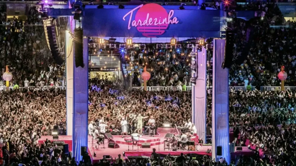 Uma grande multidão reunida em um show noturno com um palco iluminado. No centro do palco, músicos tocam instrumentos, e o nome "Tardezinha" é exibido em uma tela grande acima deles.
