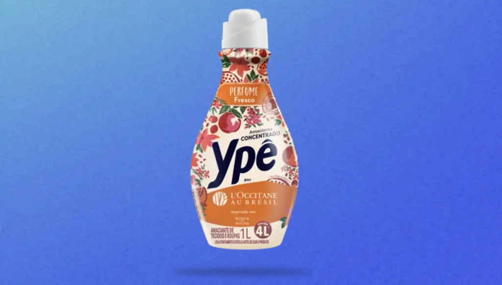 Uma garrafa de amaciante de roupas Ypê, com uma embalagem vibrante e colorida inspirada na L'Occitane au Brésil. A garrafa é predominantemente branca com um rótulo que apresenta ilustrações de folhas tropicais, flores e frutas em tons de vermelho, laranja e verde. No centro, o logotipo "Ypê" está em um círculo branco, e abaixo dele, as palavras "por L'OCCITANE AU BRÉSIL" são exibidas. Na parte superior da garrafa, há um bico branco. A garrafa está contra um fundo azul degradê. O texto na garrafa indica "PERFUME Fresco", "Amaciante CONCENTRADO", "inspirado em NINFAS ÁGUAS", e "AMACIANTE DE TECIDOS E ROUPAS 1L 4L", sugerindo que 1 litro do produto concentrado rende o equivalente a 4 litros de amaciante regular.