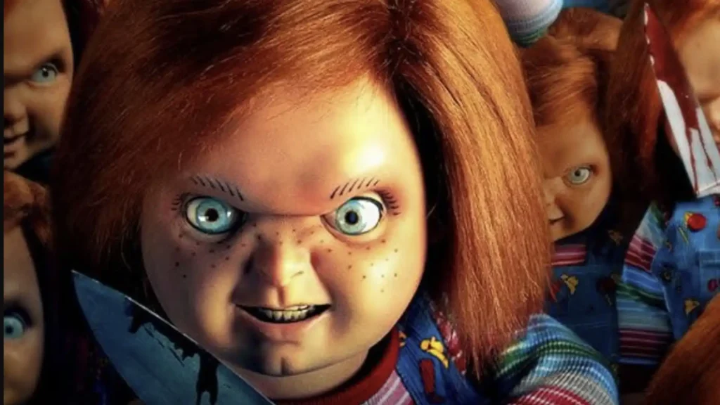 Uma imagem de close-up de um boneco Chucky de olhos azuis e cabelo ruivo, com sardas e uma expressão maliciosa. Ele está segurando uma faca. Outros bonecos Chucky são visíveis no fundo, um dos quais está ligeiramente fora de foco.