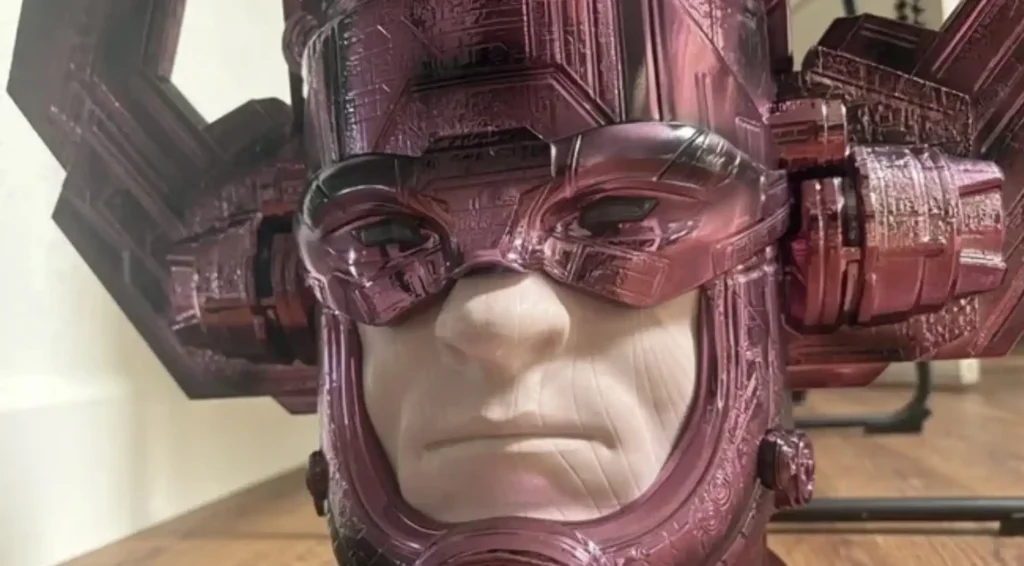 Uma imagem em close-up de um busto brilhante de Galactus, um personagem da Marvel Comics. O busto tem um capacete roxo metálico com detalhes intrincados e um rosto pálido e sério por baixo. O fundo está desfocado, mas parece ser um chão de madeira.