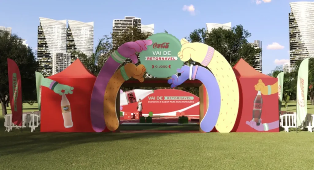 Uma entrada colorida para um evento da Coca-Cola, com um grande arco em tons pastel de roxo, amarelo e marrom claro, que se assemelham a braços humanos estilizados se encontrando no topo. No centro do arco, um círculo verde-água exibe o logotipo da Coca-Cola e o texto "VAI DE RETORNÁVEL" e "O JOGO". De cada lado do arco, há tendas vermelhas com a marca Coca-Cola. A tenda da esquerda tem uma mão verde segurando uma garrafa de Fanta, e a tenda da direita tem uma mão verde segurando uma garrafa de Coca-Cola. Ao fundo, há um palco com mais branding da Coca-Cola e o slogan "VAI DE RETORNÁVEL ECONOMIA E SABOR PARA SUAS REFEIÇÕES". O evento parece estar ocorrendo ao ar livre em um parque urbano com árvores e arranha-céus ao fundo.