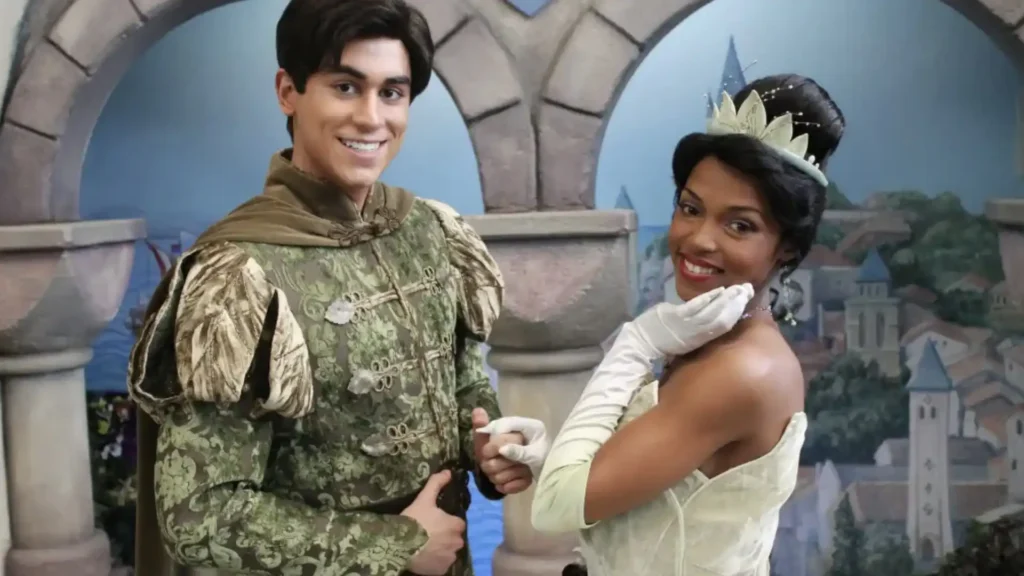 Uma imagem de um casal fantasiado de Tiana e Príncipe Naveen do filme da Disney "A Princesa e o Sapo". Eles estão em um cenário que lembra um castelo ou paisagem europeia. Ambos estão sorrindo e olhando para a câmera, com o Príncipe Naveen segurando a mão de Tiana.