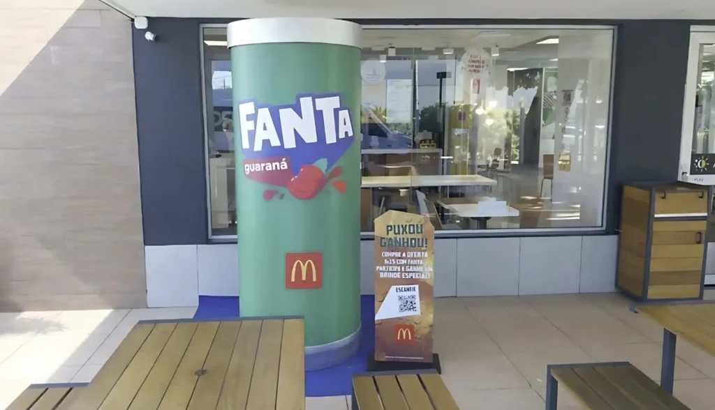 Uma lata gigante e cilíndrica de Fanta Guaraná, predominantemente verde com o logotipo da Fanta em roxo e branco e "guaraná" em branco, e o logotipo do McDonald's em vermelho e dourado, está posicionada em frente a um restaurante McDonald's. A lata está sobre uma base azul circular no chão. À direita da lata, há um totem promocional vertical que diz "PUXOU GANHOU!" no topo, com o logotipo do McDonald's e um código QR abaixo. Mesas e bancos de madeira clara estão visíveis no primeiro plano, à esquerda e à direita. O restaurante McDonald's tem grandes janelas de vidro que refletem o ambiente externo.