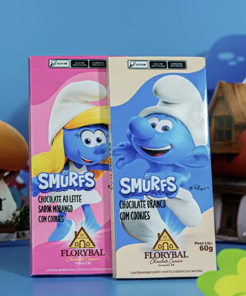 Duas barras de chocolate Florybal com tema dos Smurfs. A embalagem da esquerda é rosa, com a Smurfette sorridente e os dizeres "OS SMURFS CHOCOLATE AO LEITE SABOR MORANGO COM COOKIES". A embalagem da direita é azul, com o Papai Smurf sorridente e os dizeres "OS SMURFS CHOCOLATE BRANCO COM COOKIES". Ambas as embalagens exibem o logotipo da Florybal, indicam "ALTO EM AÇÚCAR ADICIONADO" e "GORDURA SATURADA", e "PESO LÍQ.: 60g". O fundo desfocado sugere um ambiente lúdico.