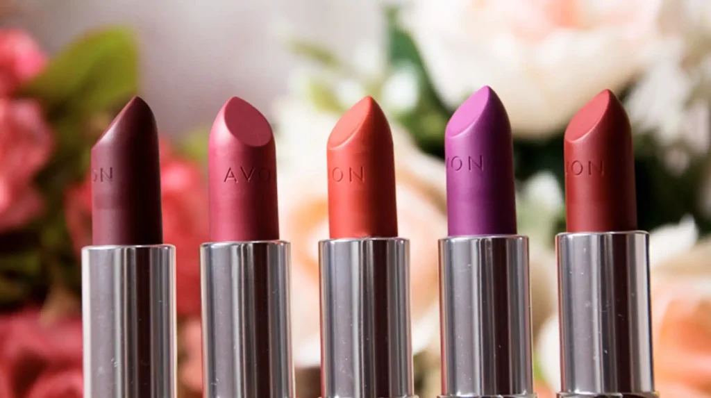 Uma fila de cinco batons da marca Avon em tons que variam do borgonha ao rosa, coral e roxo, com um fundo desfocado de flores em tons de rosa, branco e verde.