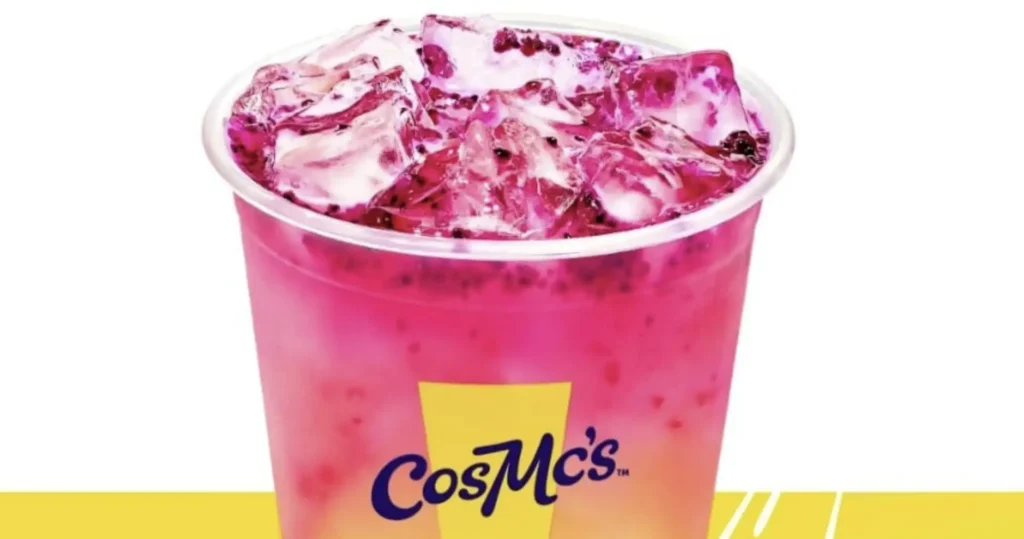 Uma foto de uma bebida CosMc's do McDonald's, que é rosa vibrante com gelo e o que parecem ser pedaços de frutas vermelhas escuras ou sementes misturadas. A bebida está em um copo transparente com o logotipo amarelo e azul "CosMc's" visível na frente. O fundo é branco na parte superior e amarelo na parte inferior.