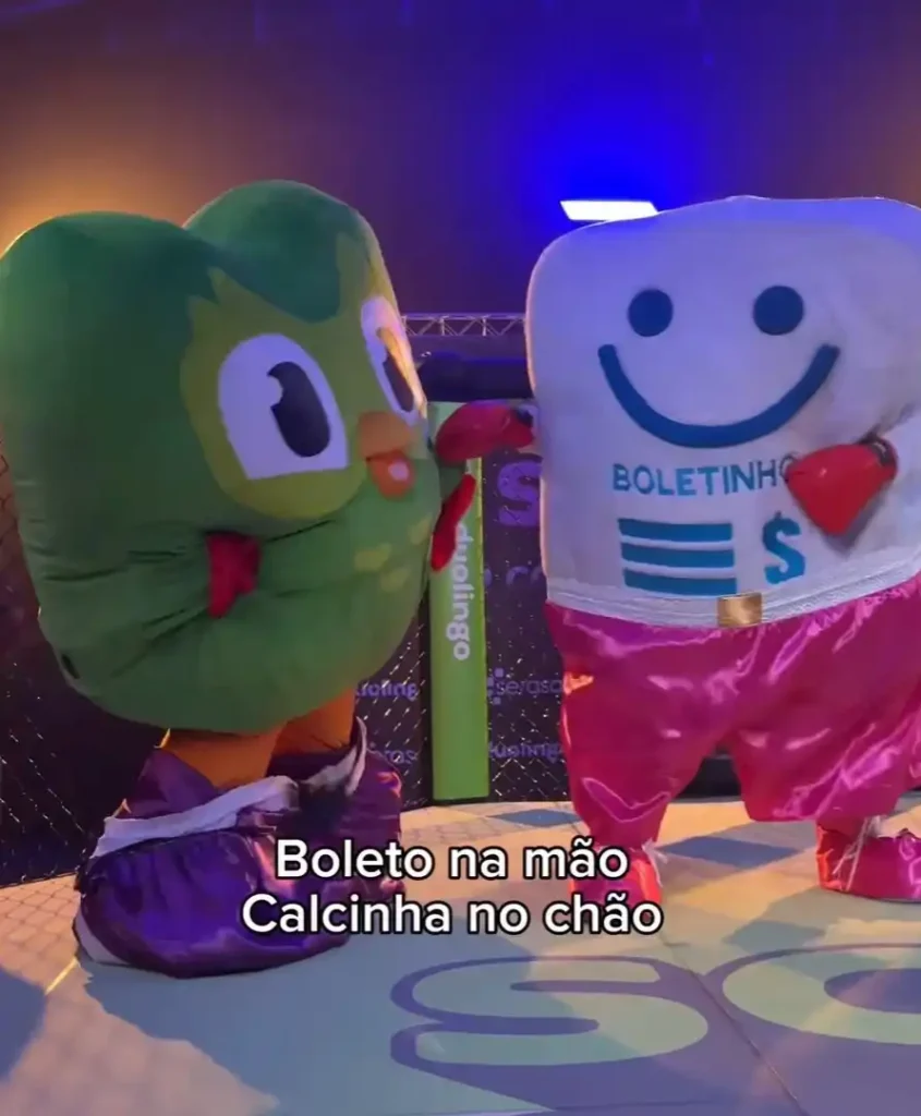 Uma imagem mostra duas mascotes de desenho animado em tamanho real. À esquerda está a mascote verde do Duolingo, um coruja chamada Duo, usando luvas de boxe vermelhas. À direita está uma mascote branca em forma de dente sorridente, também usando luvas de boxe vermelhas e shorts rosa com "BOLETINHO" e um cifrão estampado na parte superior. Ambos estão em uma área que se assemelha a um ringue de boxe ou academia. Na parte inferior da imagem, um texto em português diz "Boleto na mão Calcinha no chão".