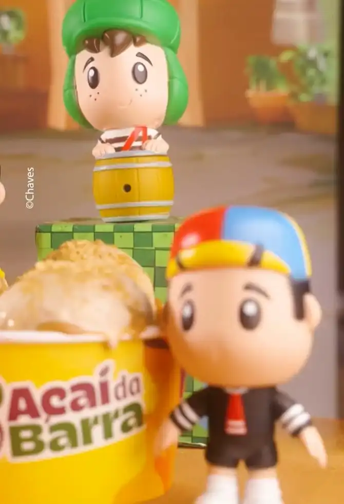 Uma colher de metal prateada está levantada sobre um copo amarelo com a inscrição "Açaí da Barra", contendo açaí e granola, enquanto um boneco estilizado do Quico de "Chaves" está ao lado direito, olhando para o lado.