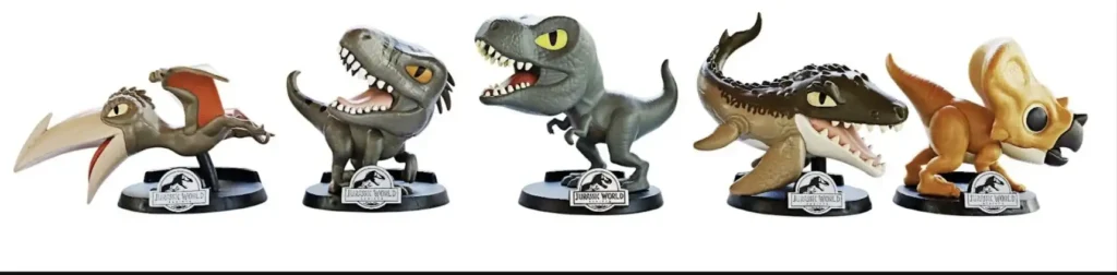Cinco brinquedos colecionáveis de dinossauros do Jurassic World, estilizados com cabeças grandes e características fofas, são exibidos em uma linha horizontal em seus pedestais pretos com o logotipo do Jurassic World. Da esquerda para a direita, os dinossauros são: um Pteranodonte marrom e bege com asas abertas; um Velociraptor azul-acinzentado com a boca aberta; um T-Rex cinza com a boca aberta; um Mosassauro marrom claro e creme com um corpo longo e dentes afiados; e um Triceratops marrom e bege.