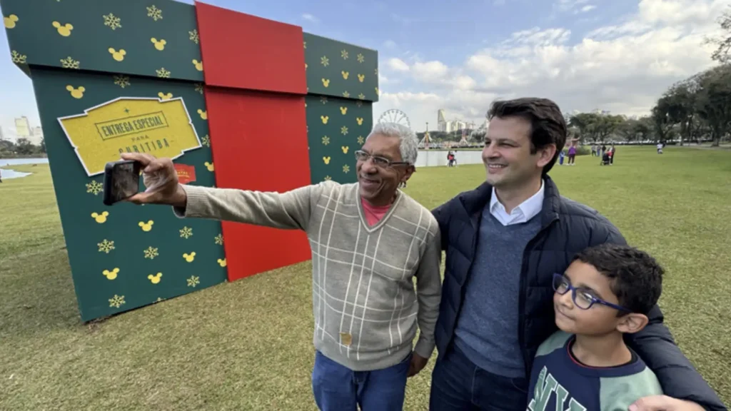 Uma grande caixa de presente verde e vermelha com desenhos de Mickey Mouse e um selo escrito "ENTREGA ESPECIAL PARA CURITIBA" ao fundo. Três pessoas posam para uma selfie em primeiro plano: um homem mais velho de camisa quadriculada cinza e branca segura um celular para tirar a foto, enquanto um homem mais jovem de jaqueta azul escura e um menino de óculos e camiseta azul escura olham para o celular sorrindo.