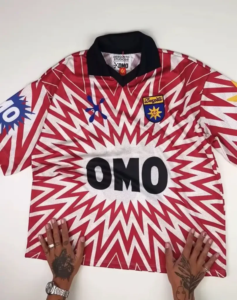 Um close-up de uma camisa de futebol branca e vermelha com o logotipo "OMO" em letras pretas arrojadas no centro, sobre uma superfície branca, com duas mãos tatuadas parcialmente visíveis nas bordas inferior, mostrando detalhes da estampa gráfica e dos emblemas da camisa.