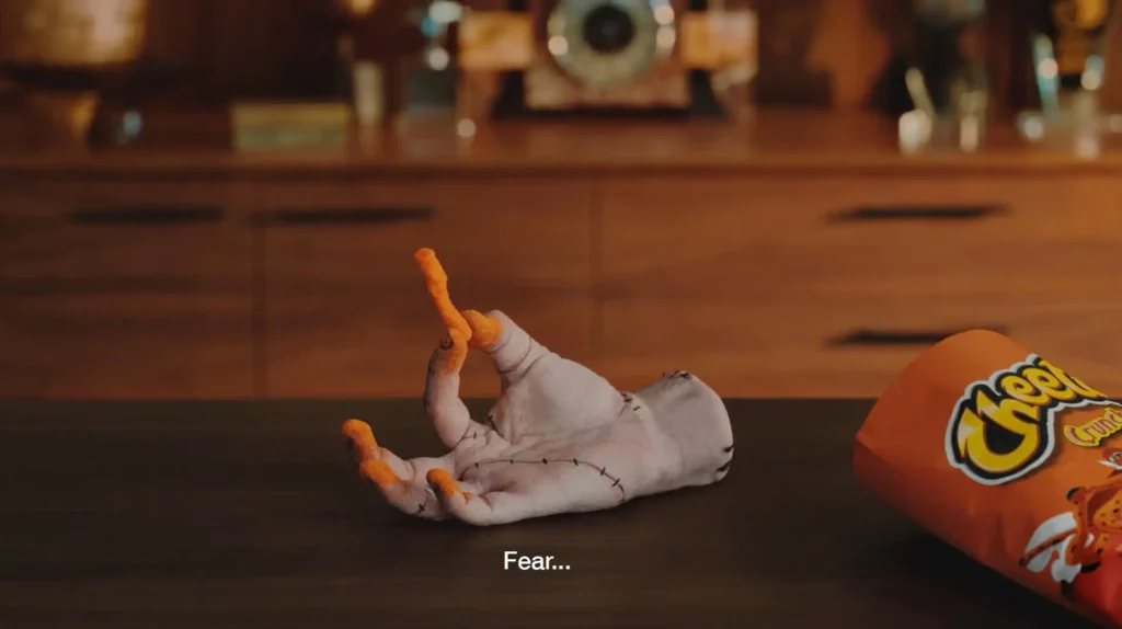 Um pacote de Cheetos Crunchy deitado ao lado de uma mão com pó laranja nos dedos, e alguns salgadinhos espalhados sobre uma superfície escura.