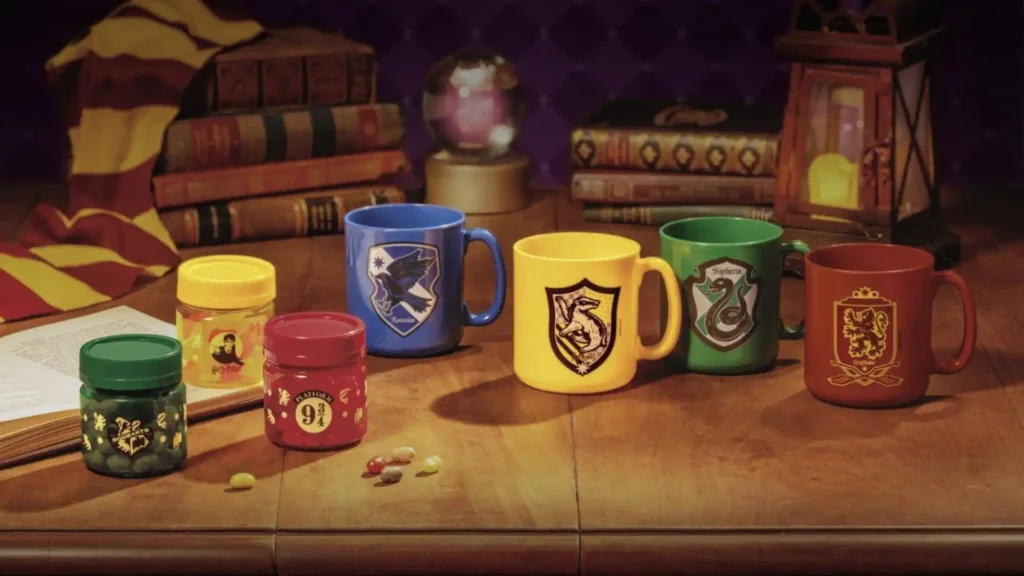 Uma imagem mostra uma mesa de madeira com vários itens temáticos de Harry Potter. Há quatro canecas coloridas, cada uma representando uma casa de Hogwarts: uma caneca azul para Corvinal, uma amarela para Lufa-Lufa, uma verde para Sonserina e uma vermelha para Grifinória. Cada caneca exibe o brasão de sua respectiva casa.
Além das canecas, há três pequenos potes de doces ou pílulas. O pote verde tem um rótulo que parece pertencer à Sonserina, o amarelo tem um rótulo com uma imagem parecida com Harry Potter, e o vermelho tem o número "9 3/4" (referente à Plataforma 9 3/4). Alguns doces estão espalhados pela mesa, perto dos potes.
Ao fundo, há pilhas de livros antigos, um globo de cristal e uma lanterna, reforçando o ambiente mágico e estudioso. Uma bandeira listrada em vermelho e dourado, cores da Grifinória, está pendurada sobre os livros no canto superior esquerdo.