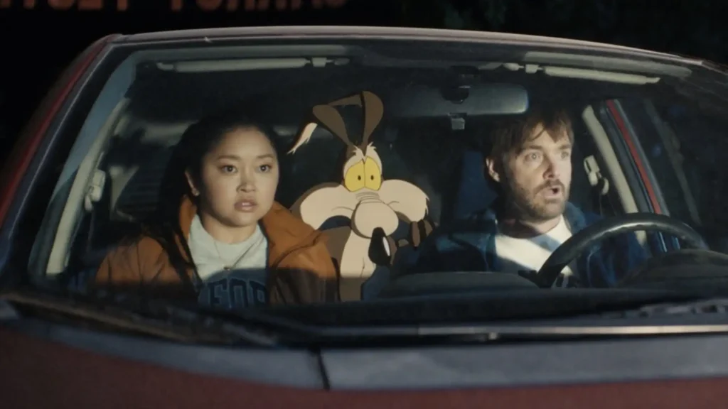 Uma imagem de uma cena de filme que mostra três personagens em um carro vermelho. À esquerda está uma jovem asiática com cabelos escuros na altura dos ombros e vestindo um casaco marrom sobre uma camiseta azul. No centro, no banco de trás, está um personagem de desenho animado, o Coiote, da Warner Bros., olhando para frente com uma expressão preocupada. À direita, ao volante, está um homem caucasiano com barba e cabelo castanho-avermelhado, usando uma camisa xadrez azul e branca. Todos os três estão olhando para frente com expressões de surpresa ou choque. O fundo através do para-brisa é escuro, com alguns borrões de luz, sugerindo que é noite ou o carro está em movimento.