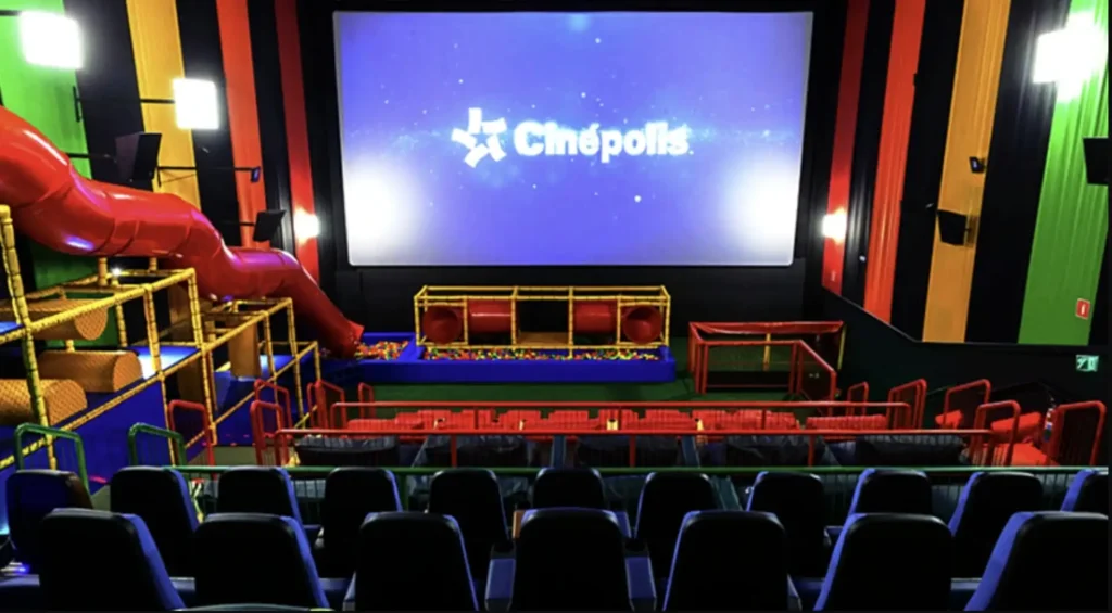 Uma sala de cinema Cinepolis Junior com escorregador vermelho, piscina de bolinhas e assentos coloridos, projetada para festas de aniversário infantis.