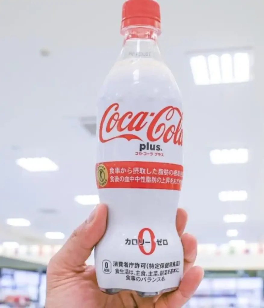 Uma garrafa branca de Coca-Cola Plus sendo segurada por uma mão. O logotipo vermelho e branco da Coca-Cola está proeminentemente exibido. Abaixo do logotipo, há um texto em japonês. Na parte inferior da garrafa, está escrito "CALORIE ZERO" em japonês e um "0" grande, indicando zero calorias. Há também um texto menor em japonês e um selo de aprovação do governo japonês para Alimentos para Uso Específico de Saúde (FOSHU). O fundo está levemente desfocado, mostrando o interior iluminado de uma loja.
