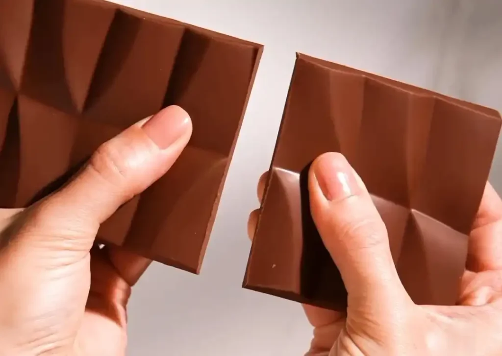 Duas mãos segurando pedaços de uma barra de chocolate de tom marrom escuro. O chocolate tem um padrão geométrico em relevo. As mãos parecem estar prestes a separar os dois pedaços.