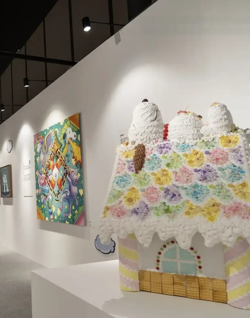 Uma foto de uma casa de gengibre ornamentada com glacê e balas coloridas em primeiro plano, ladeada por duas esculturas brancas com topos pontudos que se assemelham a cachorros de desenho animado. No fundo, uma parede de galeria branca apresenta uma pintura colorida de criaturas fantásticas e formas abstratas, iluminada por holofotes de trilha no teto. No canto inferior esquerdo, uma pequena nuvem cinza de desenho animado paira perto da base da casa de gengibre.