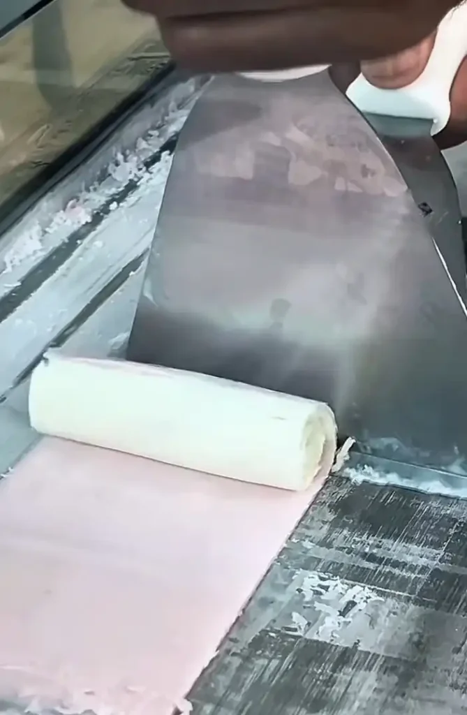 Uma mão habilidosa usa uma espátula de metal para enrolar sorvete rosa pálido que foi espalhado finamente em uma superfície gelada.