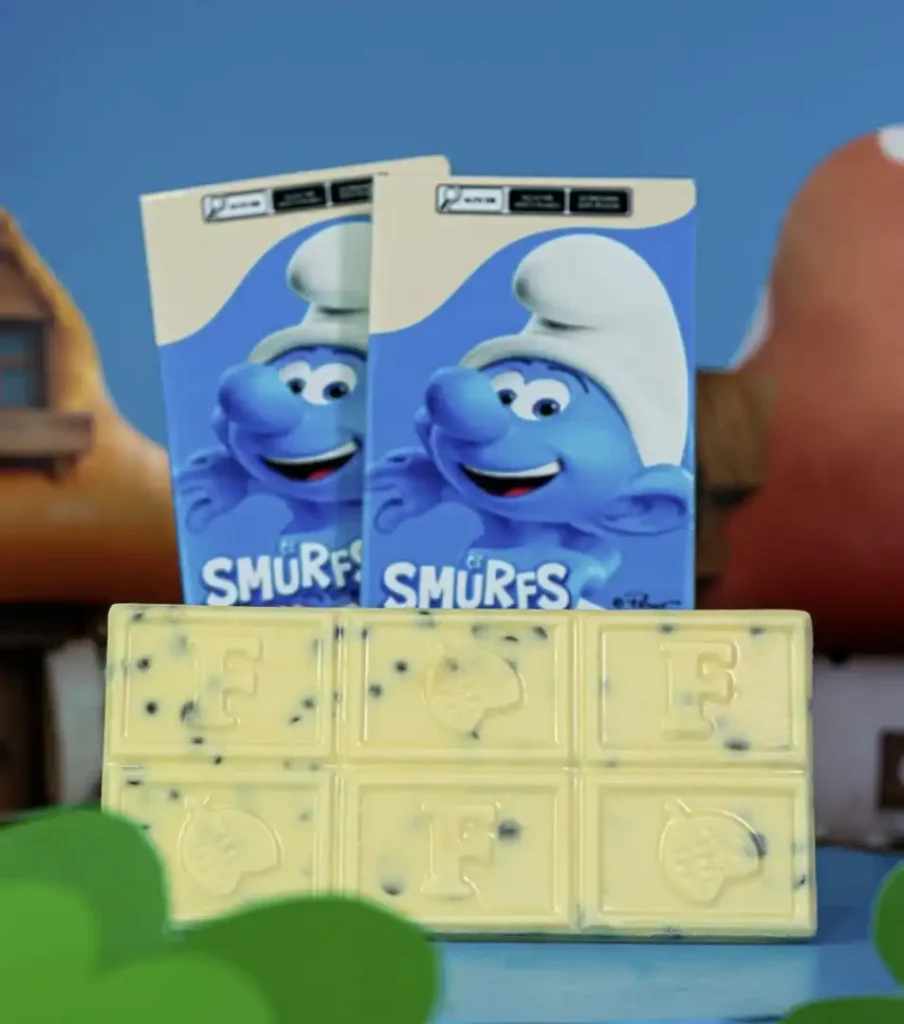 Duas barras de chocolate Florybal com tema dos Smurfs. As embalagens são azuis e exibem a imagem de um Smurf sorridente e o logotipo "SMURFS". Na frente das embalagens, há uma barra de chocolate branco dividida em seis seções. Cada seção possui uma letra em relevo: "F", "L", "O", "R", "Y", "B". As letras, no entanto, estão embaralhadas e parecem formar "F L O F R Y", com as letras "F" e "L" repetidas, e uma das seções apresenta uma bolota em relevo. O chocolate branco tem pequenas partículas escuras espalhadas, possivelmente de mirtilo ou algo semelhante. O fundo desfocado sugere um cenário de floresta ou vila.