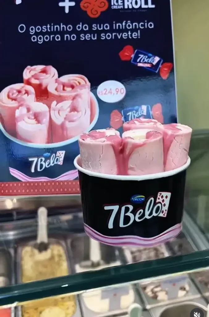 Um copo preto cheio de rolos de sorvete cor-de-rosa com calda vermelha por cima fica em uma vitrine de sorvetes. Atrás do copo, um cartaz preto anuncia "ICE CREAM ROLL" e mostra mais rolos de sorvete. O logotipo do doce 7Belo aparece no copo e no cartaz.