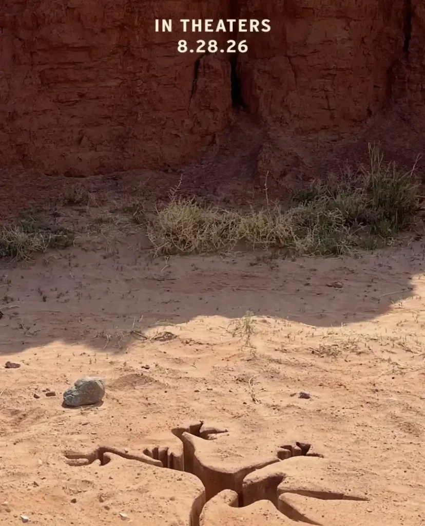 Uma imagem vertical mostra uma cena desértica com areia e formações rochosas íngremes e marrons ao fundo. No primeiro plano, na areia, há uma forma escura e irregular, que se assemelha a uma pegada de dinossauro ou a um fóssil. No canto superior central, há um texto branco que diz "IN THEATERS" e abaixo, "8.28.26".