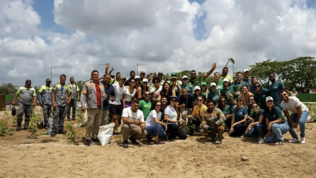 Um grupo grande e diverso de pessoas, algumas vestindo camisetas verdes e outras uniformes de trabalho, posa para uma foto em um terreno arenoso com algumas pequenas mudas plantadas. O céu é parcialmente nublado e o ambiente sugere um projeto de reflorestamento ou plantio.