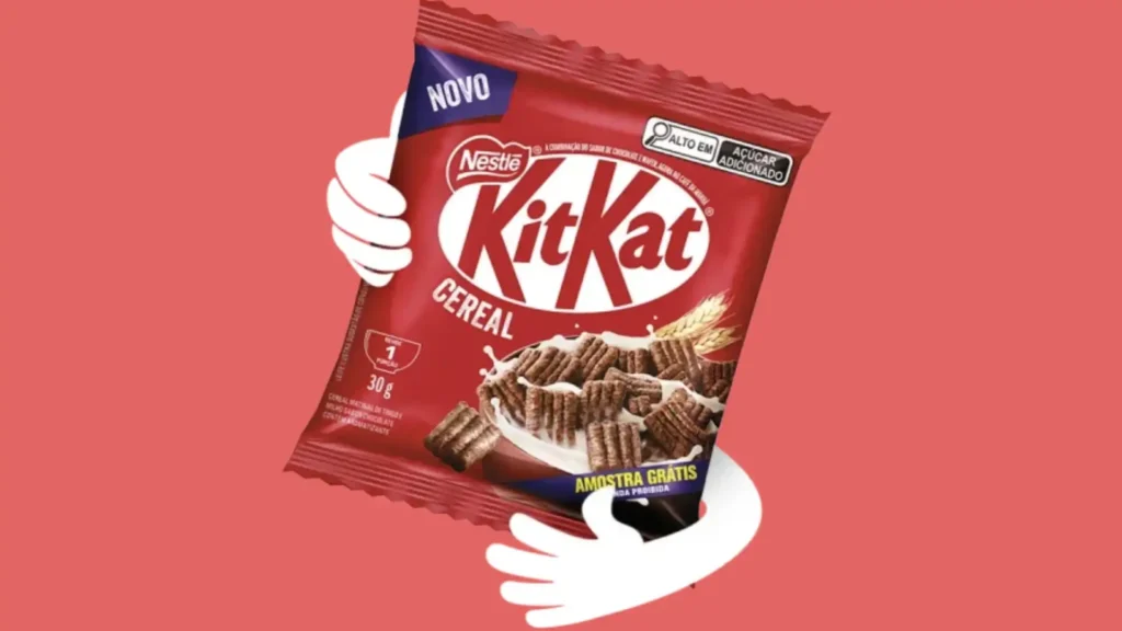 Uma sacola vermelha em forma de travesseiro de Kit Kat Cereal está centralizada em um fundo rosa claro. As palavras "Kit Kat" estão em letras brancas grandes, e a palavra "CEREAL" está em letras pretas menores. A Nestlé está logo acima das palavras "Kit Kat". Há um adesivo azul e branco no canto superior direito que diz "ALTO EM AÇÚCAR ADICIONADO". Abaixo das palavras "Kit Kat Cereal" está uma tigela branca de cereal, com pedaços de cereal Kit Kat quadrados marrons e leite branco. Algumas gotas de leite branco estão respingando para fora da tigela. À direita da tigela, há cinco talos de grãos de trigo dourados. À esquerda da sacola, há um adesivo branco em forma de elipse com um "1" preto no meio. Abaixo dele, há um "porção" cinza, um "30g" preto e um "CEREAL MATINAL COM SABOR ARTIFICIAL CHOCOLATE". No canto inferior direito da sacola está um adesivo amarelo que diz "AMOSTRA GRÁTIS" em letras azuis e "VENDA PROIBIDA" em letras menores. Duas mãos brancas, uma à esquerda e outra à direita, parecem estar segurando a sacola. As mãos se estendem de cada lado de trás da sacola.
