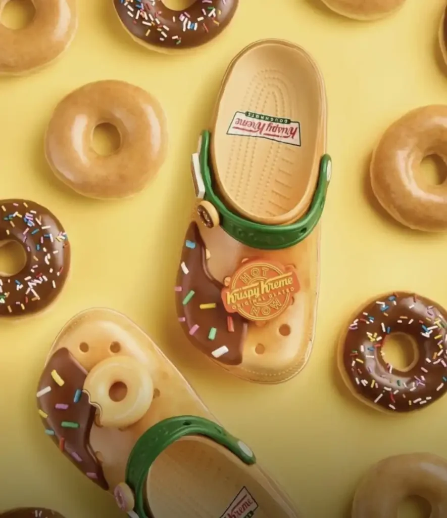 Um par de sandálias Crocs bege com detalhes de glacê de chocolate e granulado colorido, semelhantes a um donut. Um dos Crocs tem um pequeno donut mordido como enfeite. As sandálias estão cercadas por vários donuts, alguns com glacê simples e outros com glacê de chocolate e granulado, todos dispostos sobre um fundo amarelo. A marca Krispy Kreme Doughnuts é visível no interior dos Crocs.