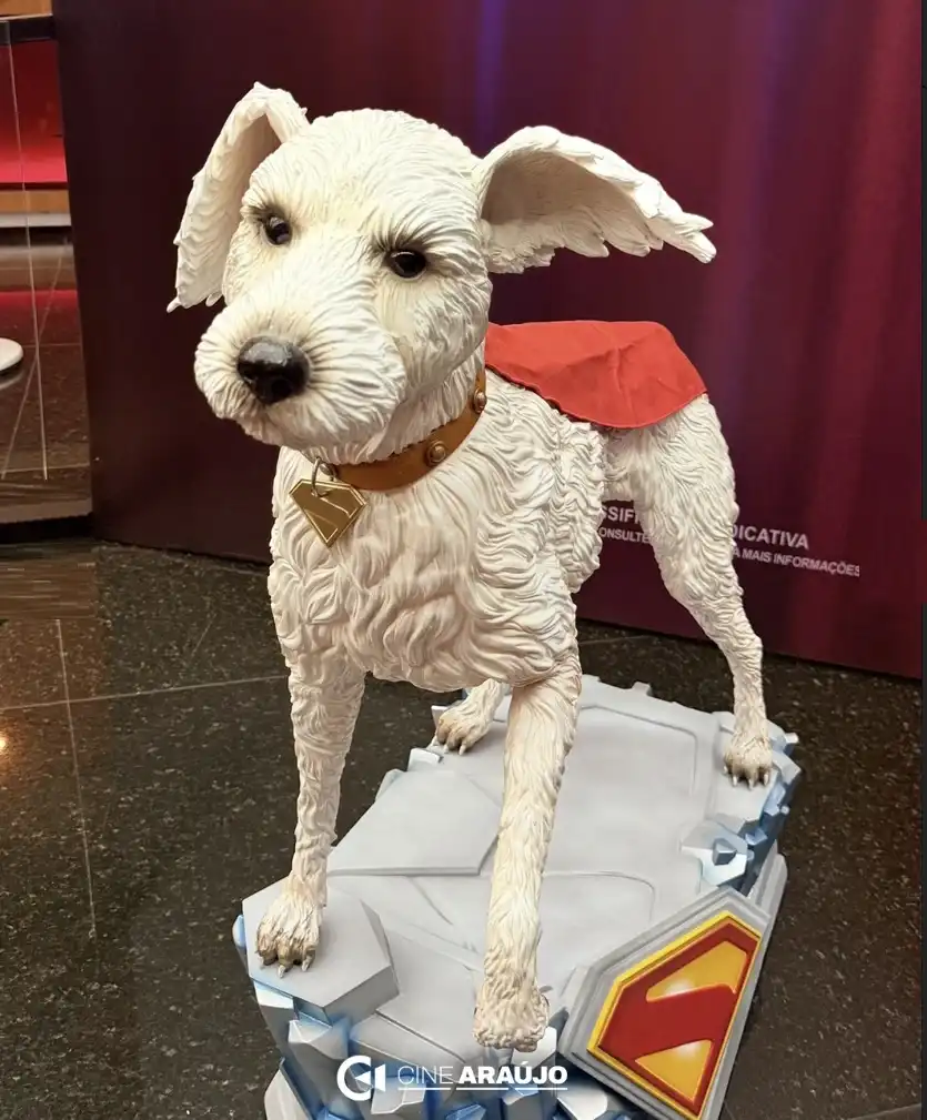 Uma estátua de Krypto, o cachorro do Superman, retratado como um cão branco com pelo texturizado, vestindo uma pequena capa vermelha e uma coleira marrom com um emblema dourado em forma de "S". Ele está sobre uma base cinza que se assemelha a cristais de gelo, com o logo do Superman na frente.