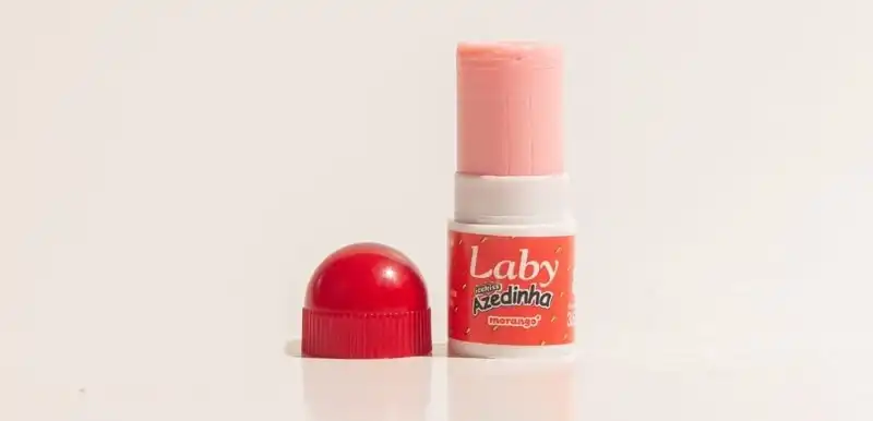 Uma imagem em close-up de um protetor labial da marca "Laby" com sabor de morango. O protetor labial rosa claro está parcialmente exposto, com a tampa vermelha translúcida removida e colocada ao lado. O fundo é branco, destacando o produto.