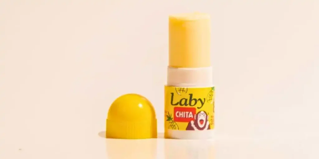 Uma imagem em close-up de um protetor labial da marca "Laby" com sabor de banana. O protetor labial amarelo está parcialmente exposto, com a tampa amarela removida e colocada ao lado. O fundo é branco, destacando o produto.
