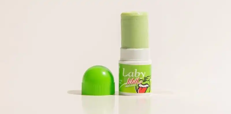 Uma imagem em close-up de um protetor labial da marca "Laby Lilith" com sabor de maçã verde. O protetor labial verde claro está parcialmente exposto, com a tampa verde brilhante removida e colocada ao lado. O fundo é branco, destacando o produto.