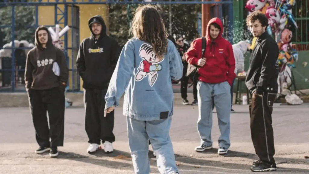 Uma imagem mostra cinco jovens, três homens e duas mulheres, em um ambiente externo que parece ser uma rua ou praça. Quatro dos jovens estão de frente, olhando para a quinta pessoa, uma mulher de cabelo longo e encaracolado que está de costas para a câmera.
A mulher de costas usa um conjunto de jeans claro, composto por uma jaqueta e calça folgada. Na parte de trás da jaqueta, há um desenho grande de um personagem de desenho animado, que se assemelha à Mônica da Turma da Mônica, segurando um skate. Seus braços estão levemente levantados e as mãos abertas, como se estivesse se apresentando ou interagindo com os outros.
Os homens e a outra mulher usam roupas folgadas de estilo streetwear, incluindo moletons, calças largas e tênis. Um homem à esquerda veste um moletom marrom e calça preta. Outro, ao lado dele, usa um moletom preto e calça preta. O homem à direita veste um moletão vermelho e calça jeans clara. E a mulher na extrema direita, veste um casaco preto e calça marrom.
No fundo, é possível ver algumas estruturas urbanas, como cercas, árvores e, à direita, balões coloridos, sugerindo um parque ou área de lazer. A luz ambiente indica ser dia.