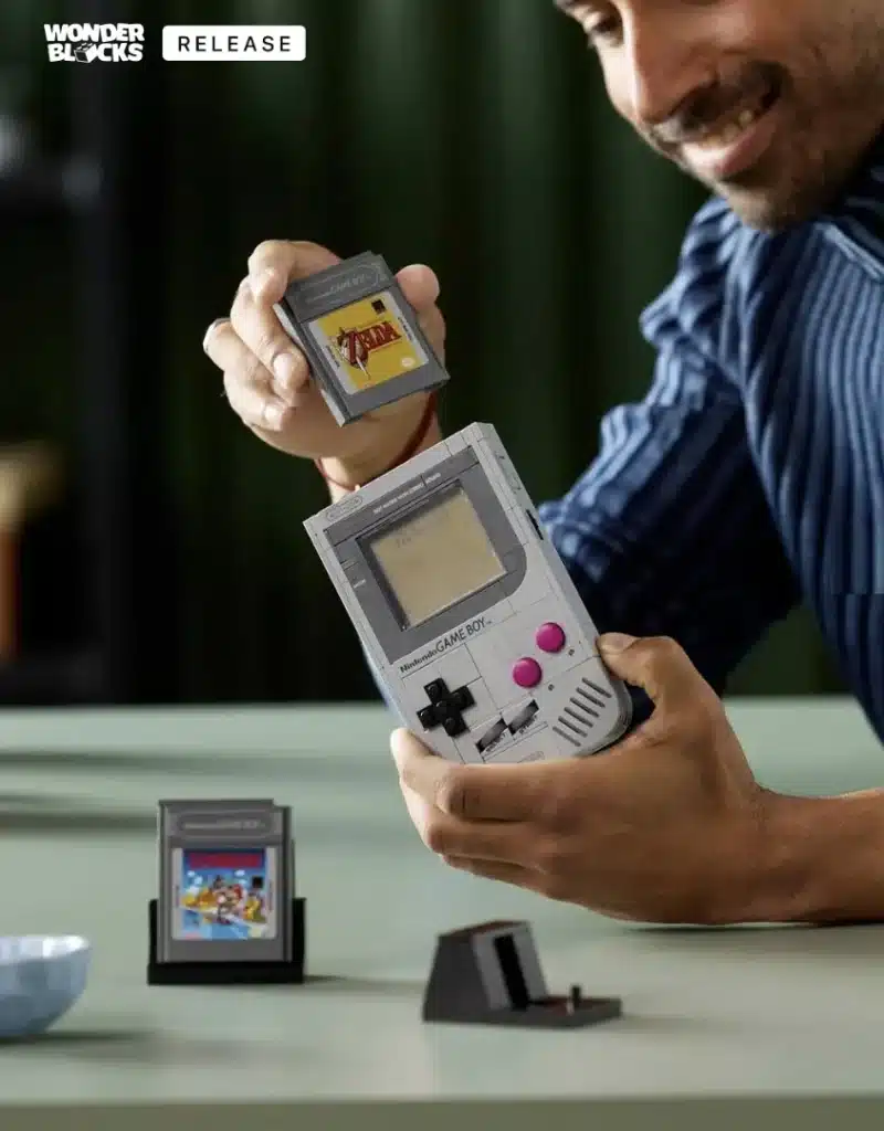 Um homem sorridente segura um Game Boy em uma mão e um cartucho de jogo na outra, prestes a inseri-lo. Há um segundo cartucho de jogo em um suporte na mesa e outro suporte vazio ao lado. No canto superior esquerdo da imagem, há o logo "WONDER BLOCKS" e a palavra "RELEASE".