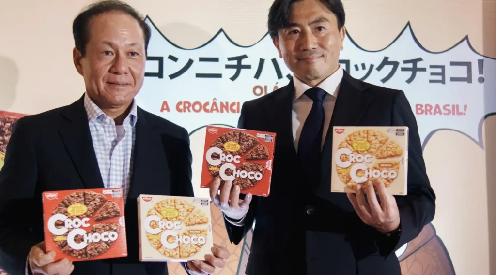 Duas pessoas sorridentes, possivelmente executivos da Nissin, seguram caixas de "Croc Choco". As caixas contêm biscoitos em formato de pizza, alguns com cobertura de chocolate escuro e outros com cobertura branca. O fundo apresenta um banner com texto em japonês e português, incluindo as frases "Olá Brasil!" e "A crocância".