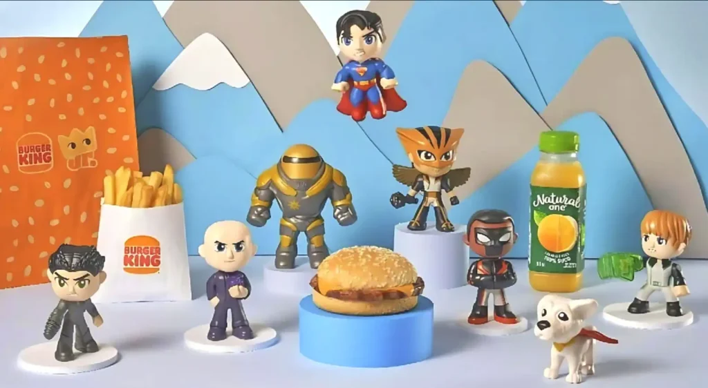 Uma imagem promocional dos brindes do Burger King. No centro, sobre um pedestal azul, há um hambúrguer. Ao redor, em um cenário que simula montanhas azuis e brancas ao fundo, com um padrão laranja no lado esquerdo, estão diversos bonecos de super-heróis em estilo "cabeçudos" ou chibi, além de um cachorro branco com uma capa vermelha. Entre os brindes, há também uma embalagem de batatas fritas do Burger King, uma sacola de papel da marca e uma garrafa de suco "Natural One" de laranja. Os personagens incluem Superman, Gavião Negro, e outros que parecem ser personagens da DC Comics, alguns em poses de ação.