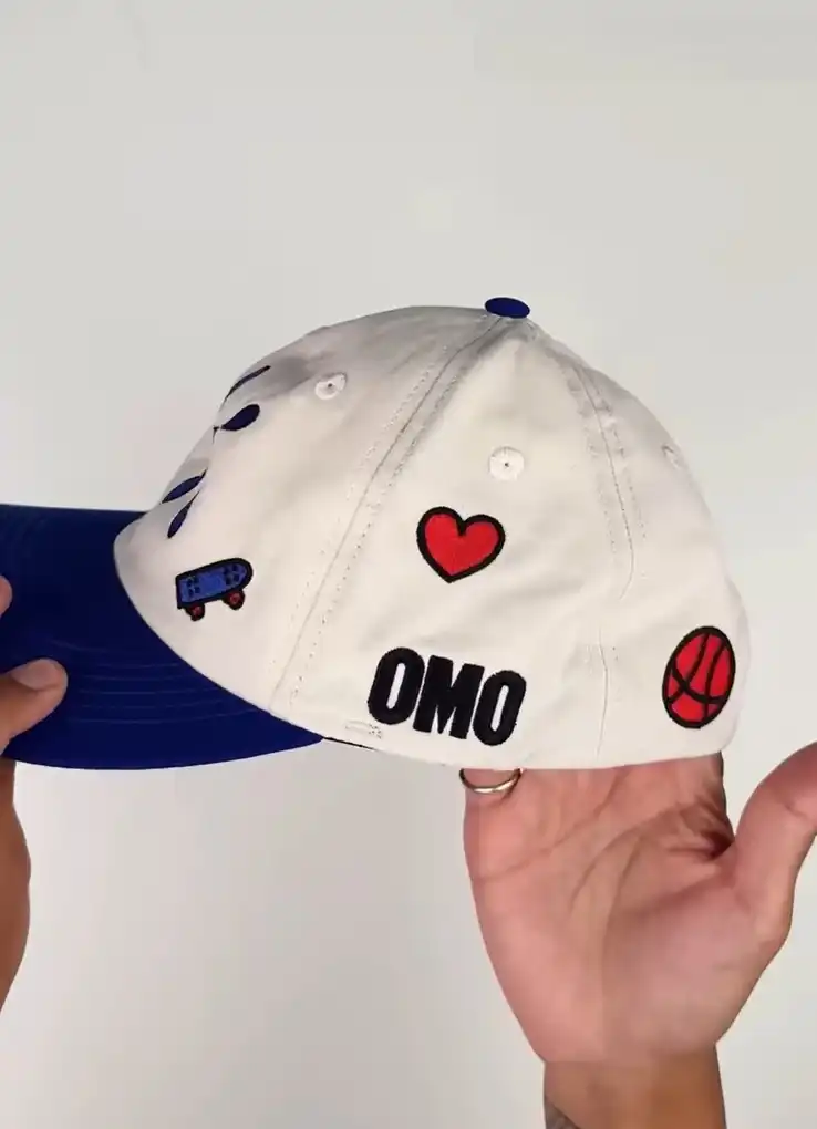 Um boné de beisebol com painéis frontais e laterais brancos e uma aba azul. O boné é decorado com vários adesivos ou patches. Na parte de trás, um logotipo "OMO" preto é visível. Outros adesivos incluem um coração vermelho, uma bola de basquete vermelha com detalhes em preto e um skate.