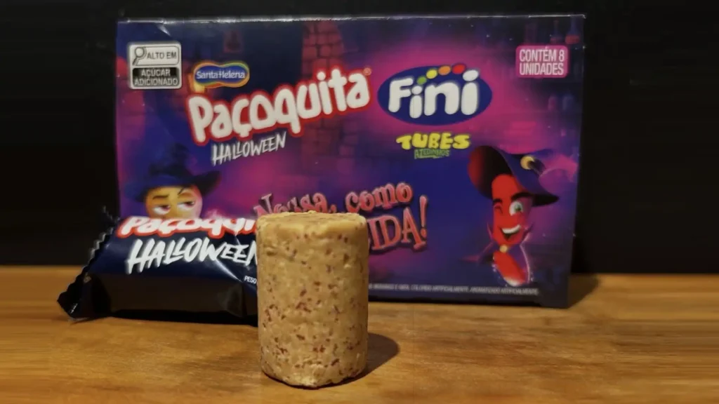 Uma caixa roxa e preta de "Paçoquita Fini Halloween" é exibida em uma superfície de madeira. A caixa mostra um personagem de desenho animado usando um chapéu de bruxa e segurando uma abóbora, e um pimentão de desenho animado usando um chapéu de bruxa. Na frente da caixa, um texto diz "Nossa, como arrepia!". Na frente da caixa, no canto superior direito, um pequeno texto branco diz "CONTÉM 8 UNIDADES". Na frente da caixa, no canto superior esquerdo, um pequeno texto branco diz "ALTO EM AÇÚCAR ADICIONADO". Ao lado da caixa, há uma paçoca cilíndrica. Uma embalagem azul escura de "Paçoquita Halloween" está à esquerda da paçoca.