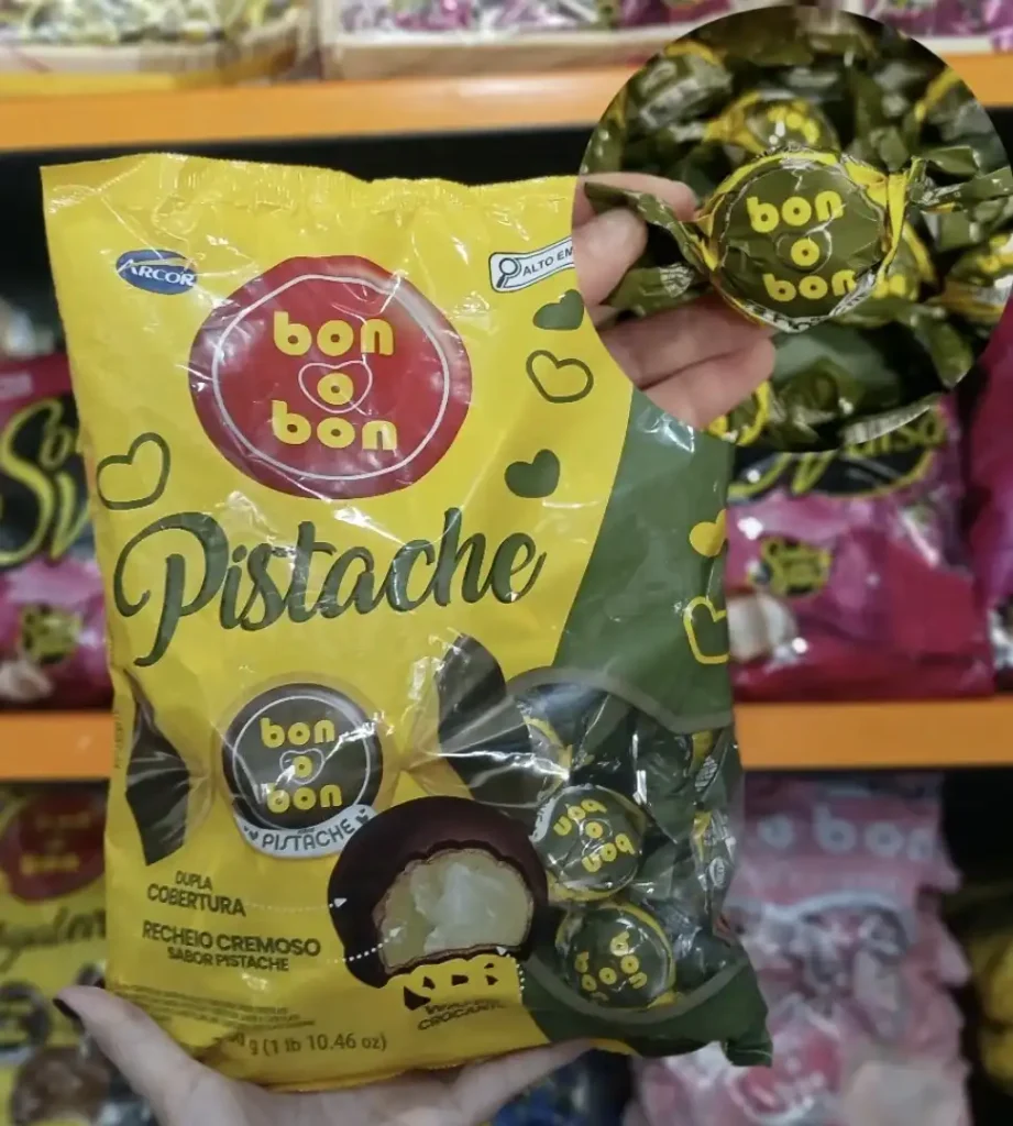 Uma pessoa segura uma embalagem amarela e verde de doces Bon o Bon sabor pistache da Arcor. A embalagem mostra bombons individuais e um bombom cortado ao meio, revelando o recheio cremoso de pistache e uma dupla cobertura. Há um círculo ampliado no canto superior direito mostrando de perto um dos bombons embalados individualmente.