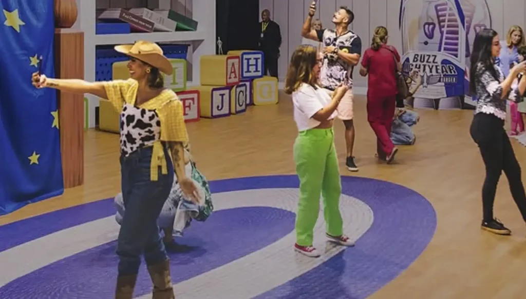Uma mulher vestida como Jessie, a cowgirl de Toy Story, posa para uma foto em um evento temático da Pixar. Ela está no centro-esquerda da imagem, com um chapéu de cowboy marrom, camisa xadrez amarela e branca, um colete estampado de vaca e jeans azul. Ela estica o braço esquerdo para tirar uma selfie com um telefone que não está visível. Outras pessoas estão ao redor em segundo plano, algumas tirando fotos ou filmando. O chão é azul com um grande círculo branco e listras azuis. Ao fundo, há cubos grandes com letras "A", "H", "J" e "C", e uma representação do Buzz Lightyear.