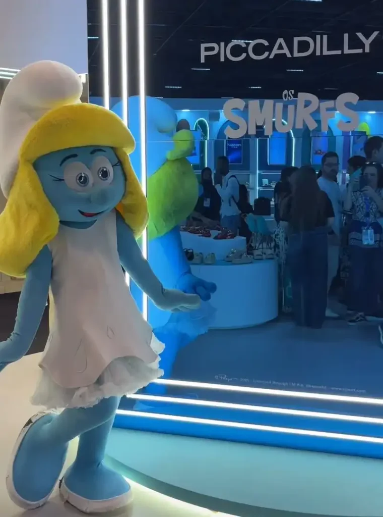 Uma figura fantasiada de Smurfette, a Smurf feminina com cabelo loiro e um vestido branco, posa em pé. Ao fundo, uma placa branca com letras maiúsculas pretas lê "PICCADILLY" e, abaixo, letras azuis maiores soletram "OS SMURFS". Atrás da Smurfette, há uma vitrine azul iluminada e algumas pessoas borradas.