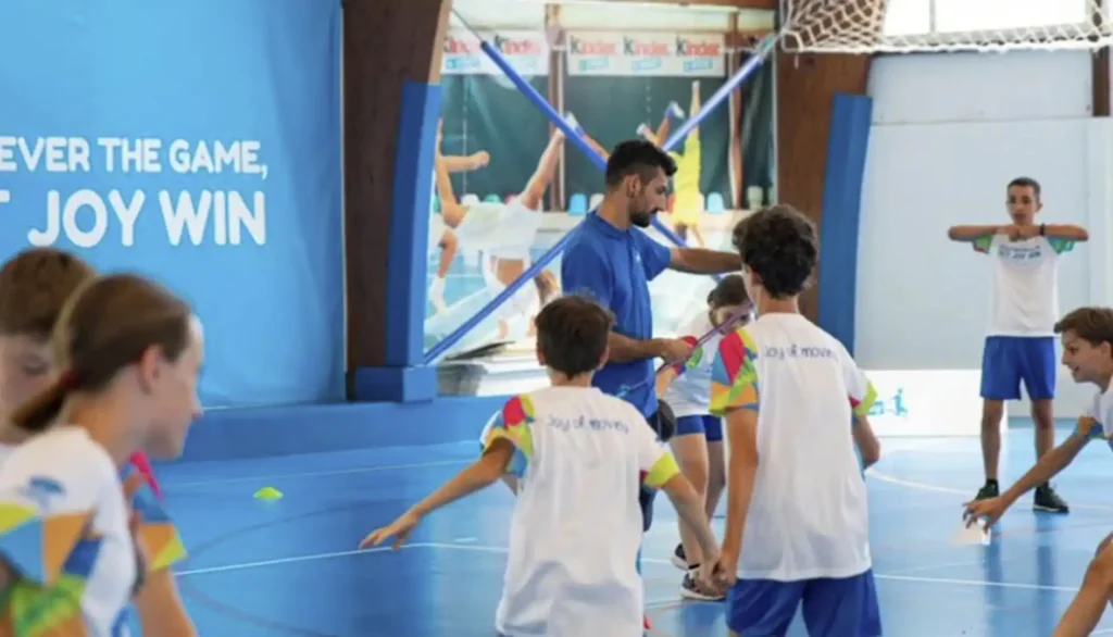 Um grupo de crianças com camisetas brancas e estampas coloridas está brincando em uma quadra poliesportiva. Um homem adulto, vestindo uma camiseta azul e segurando um bastão, interage com elas no centro da imagem. Ao fundo, uma parede azul exibe a frase "VER THE GAME, JOY WIN" em letras brancas. Outro adulto, de camiseta branca e bermuda azul, observa as crianças à direita.