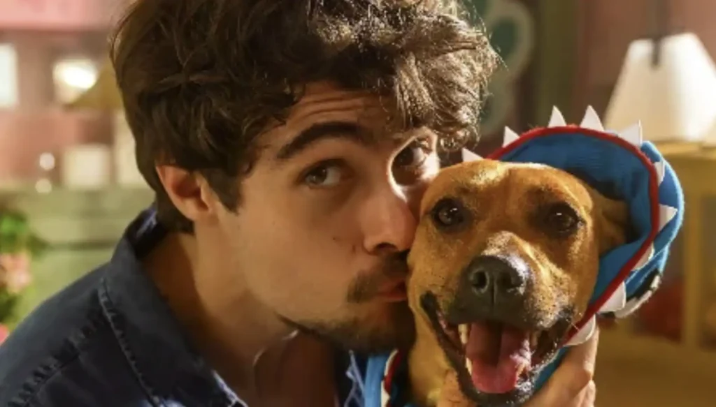 Um homem de cabelos escuros e ondulados, com uma barba rala, inclina-se para beijar um cachorro caramelo. O cachorro, que parece sorrir, usa uma fantasia azul e vermelha com barbatanas pontudas, lembrando um tubarão. A cena é bem iluminada, com um fundo desfocado