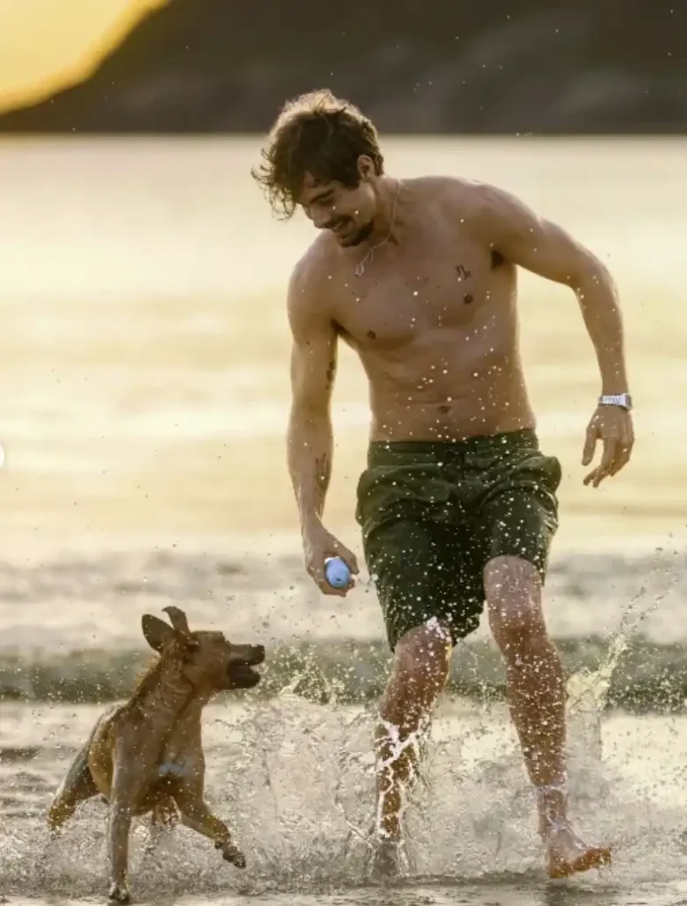 Um homem sem camisa, com shorts verdes, sorri enquanto brinca com um cachorro caramelo na água, com salpicos e reflexos do sol ao fundo.