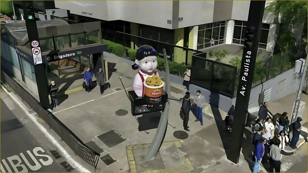 Uma estátua gigante da boneca de "Round 6" ("Squid Game") está posicionada na esquina da Avenida Paulista com a Brigadeiro em São Paulo, ao lado da estação de metrô Brigadeiro. A boneca está segurando um balde de nuggets da Seara. Pessoas estão caminhando ao redor da estátua e na calçada movimentada. O dia parece estar claro.