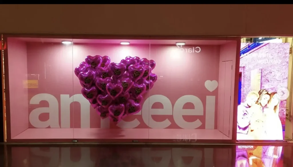 Uma vitrine rosa pastel exibe um aglomerado de balões roxos brilhantes em forma de coração no centro. Atrás dos balões, letras grandes e brancas soletram "ameei". No lado direito da vitrine, uma tela digital mostra quatro pessoas em uma foto em tons sépia.